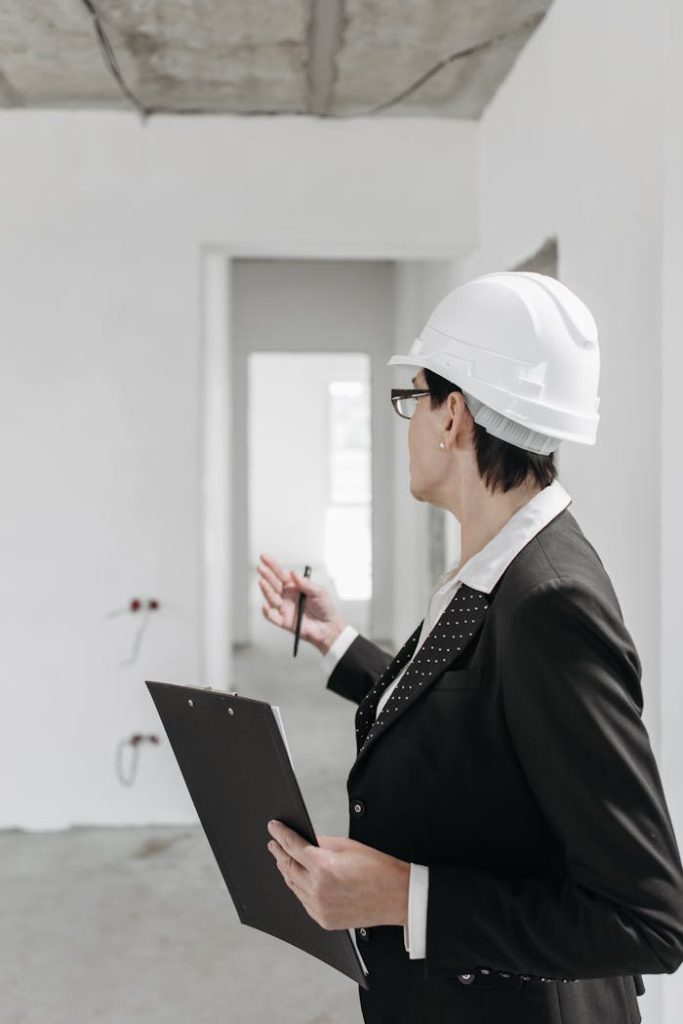 female-engineer-in-an-unfurnished-room-7937360 A professional engineer in a suit and hard hat inspecting a construction site with a clipboard.